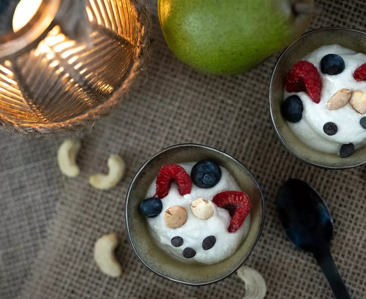 Deux ramequins noirs de crème de cajou et poires, garnis de framboises, myrtilles, amandes et pépites de chocolat, vus du dessus