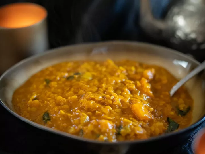 Bol de dahl de lentilles rouges fumant, devant une théière métallique et une bougie, sur un tissu sombre