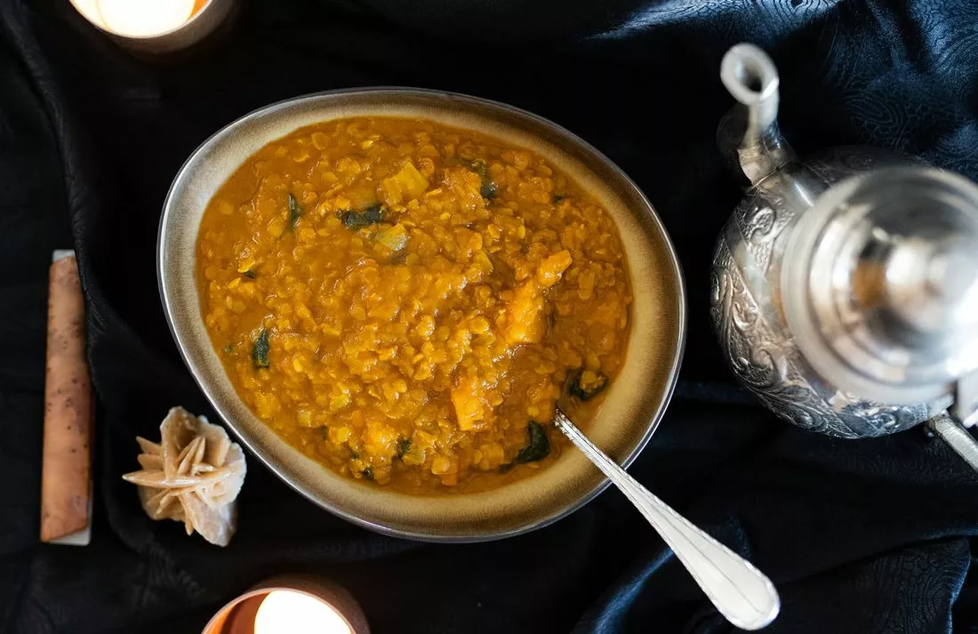 Dahl de lentilles rouges vu du dessus dans un bol ovale, avec une cuillère, une théière métallique et des bougies sur un tissu bleu nuit