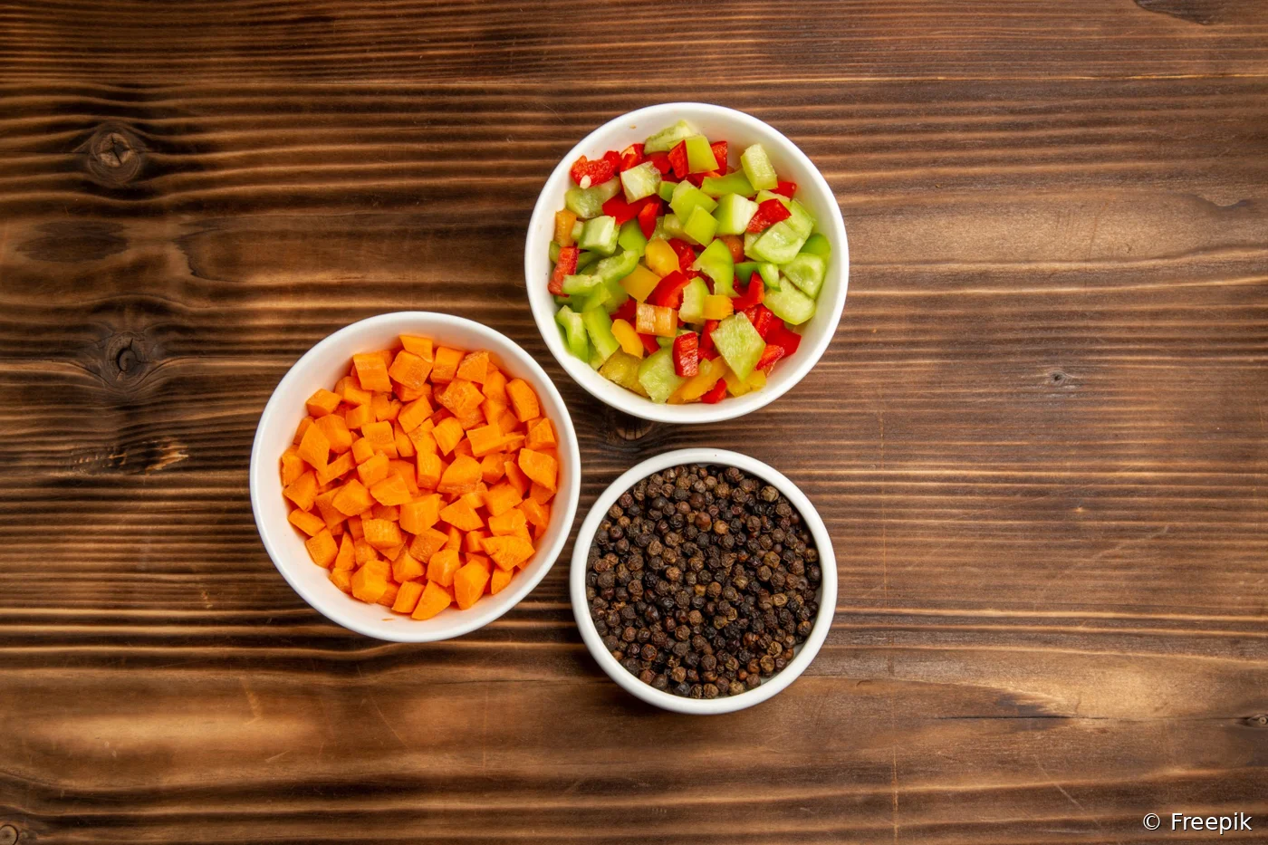 Petits dés de carotte et de poivron rouge, vert et jaune dans des bols blancs, à côté d'un bol de grains de poivre, sur une table en bois
