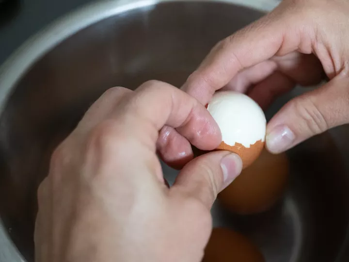 Deux mains qui écalent délicatement un œuf dur au-dessus d'un saladier en inox