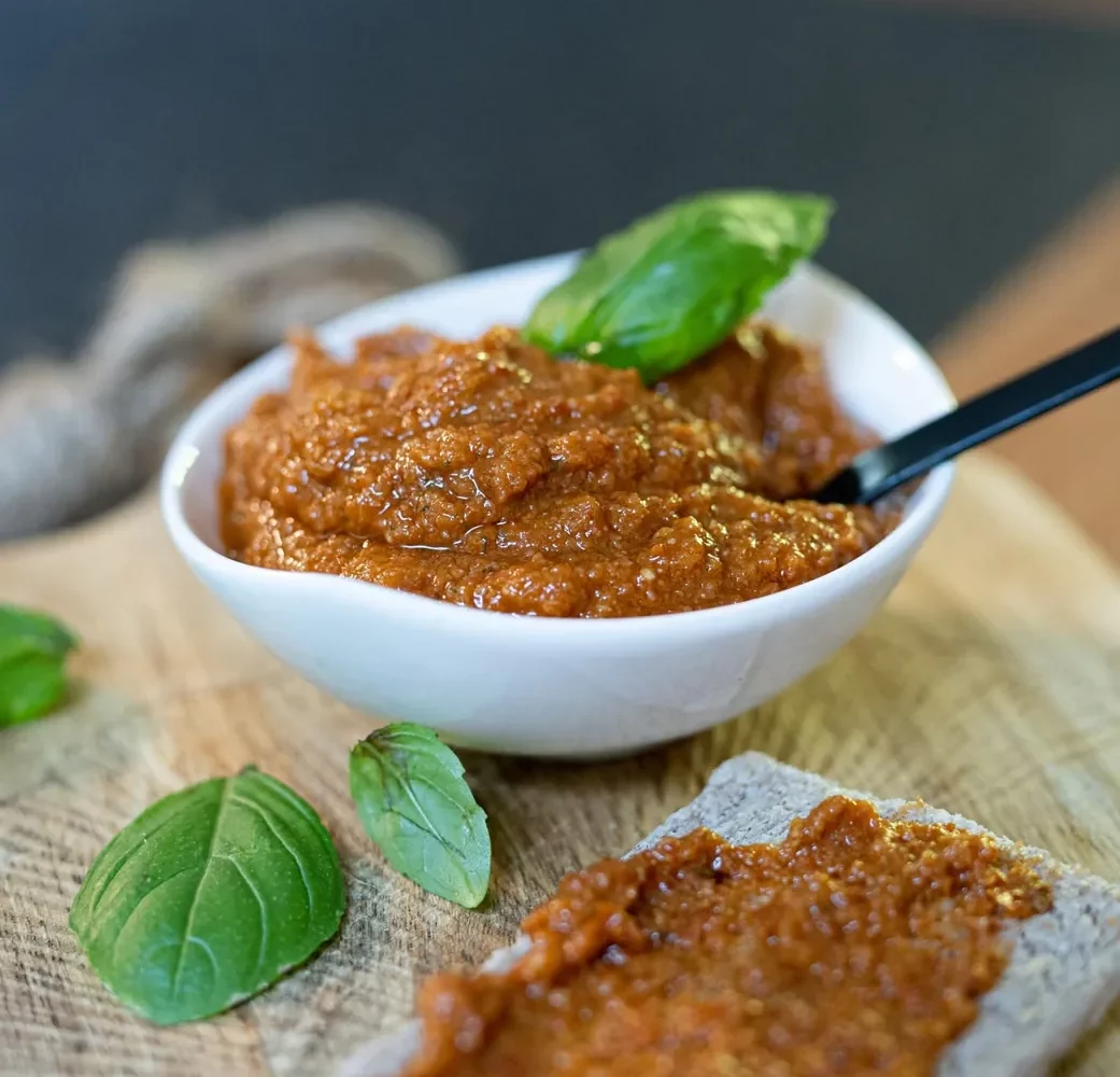 Pesto rosso maison dans un petit bol blanc, feuilles de basilic et tartine de pain