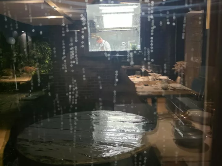Vue sur la cuisine ouverte de Luma depuis la salle, à travers une vitre couverte de gouttes de pluie
