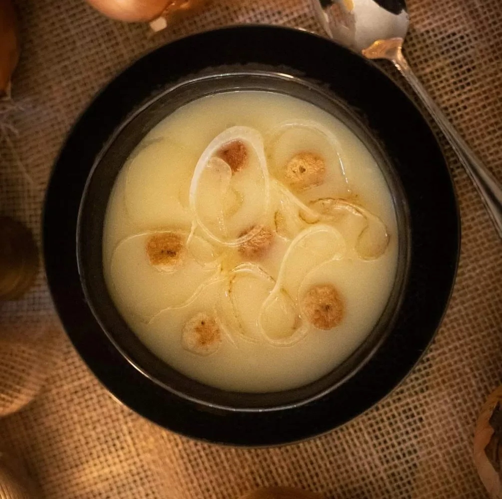 Soupe aux oignons dans un bol noir, garnie de rondelles d'oignon et de petits croûtons dorés, posée sur une toile de jute avec une cuillère et des oignons